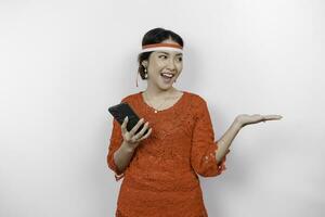 A happy Asian woman wearing red kebaya and headband, holding her phone, and pointing copy space beside her, isolated by white background. Indonesia's independence day photo
