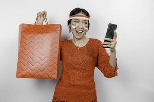 An excited Asian woman wearing red kebaya holding a shopping bag and her smartphone, studio shot isolated on white background. Indonesia's Independence day concept photo