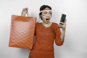 A shocked Asian woman wearing red kebaya holding a shopping bag and her smartphone, studio shot isolated on white background. Indonesia's Independence day concept photo