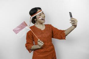 A happy Asian woman wearing red kebaya and headband, holding her phone and Indonesia's flag, isolated by white background. Indonesia's independence day photo