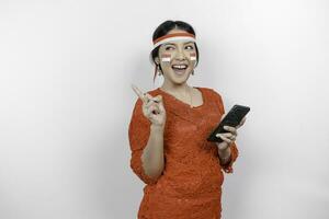 A happy Asian woman wearing red kebaya and headband, holding her phone, and pointing copy space on top of her, isolated by white background. Indonesia's independence day photo