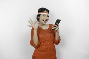 A cheerful Asian woman wearing red kebaya and headband, holding her phone while gesturing OK sign with her fingers, isolated by white background. Indonesia's independence day concept photo