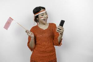 A happy Asian woman wearing red kebaya and headband, holding her phone and Indonesia's flag, isolated by white background. Indonesia's independence day photo