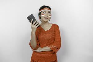 A thoughtful Asian woman wearing red kebaya and headband, holding her phone and stomach, thinking about what to eat or order, isolated by white background. Indonesia's independence day photo