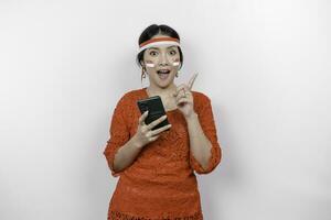 un contento asiático mujer vistiendo rojo kebaya y venda, participación su teléfono, y señalando Copiar espacio en parte superior de su, aislado por blanco antecedentes. de indonesia independencia día foto