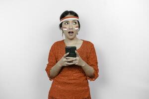 A smart young woman wearing headband and red kebaya is looking aside to an idea on copy space while holding her phone, isolated by white background. Indonesia's independence day. photo