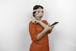 A portrait of a shocked Asian woman wearing red kebaya and headband, holding her phone, and pointing copy space on top of her, isolated by white background. Indonesia's independence day concept photo