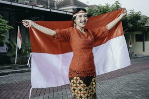 Happy smiling Indonesian woman wearing red kebaya holding Indonesia's flag to celebrate Indonesia Independence Day. Outdoor photoshoot concept photo