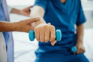 asiático fisioterapeuta Ayudar masculino paciente extensión brazo durante ejercicio correcto con pesa en mano durante formación mano en hospital foto