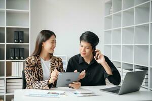 Asian business colleague business presents and explains and using laptop and tablet. Teamwork, financial marketing team,   while sitting in modern office room. photo