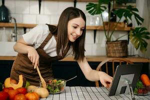 joven mujer Cocinando cena tiene vídeo llamada conversacion en cocina. sonriente contento hembra hablando con amigo utilizando solicitud en computadora. foto