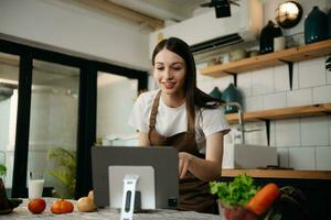 Young Woman cooking dinner has video call conversation in kitchen. Smiling happy female talking with friend using application on computer. photo