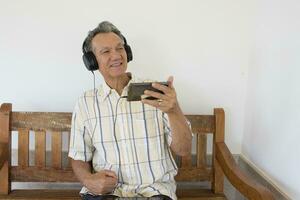 Mature Man outside sitting on a bench watching a show on his Smart Phone wearing headphones photo