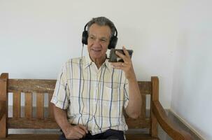 Mature Man outside sitting on a bench watching a show on his Smart Phone wearing headphones photo