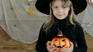 un pequeño niña en un bruja sombrero participación un calabaza video