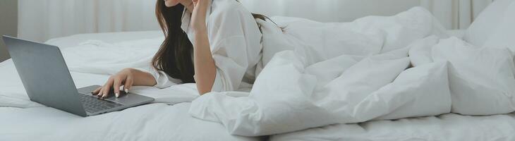 Happy young female in casual clothes smiling and looking at camera while using laptop on bed in bedroom at home photo