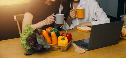 Happy two young women looking laptop computer during cooking together in kitchen room at home. Two young diverse lesbian women spending time together. LGBT and gender identity concept photo