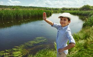 Smiling cute boy waving hello, standing on the background of the river bank. Child boy on summer vacation in the countryside photo