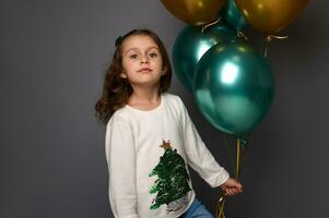 Adorable 4 years little European girl wearing white sweater with Christmas tree, holds beautiful golden and green metallic air balloons in her hand, poses against gray background looking at camera photo