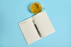 plano laico de un abierto bloc con palabra querido diario y un tinta bolígrafo siguiente a un té taza en azul antecedentes con Copiar espacio foto