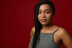 Close-up portrait of a charming young African woman with stylish traditional dreadlocks, cute smiling looking at camera, isolated over red background with copy space photo