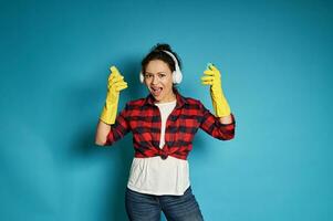 Young african woman in red plaid shirt with headphones on her head sings, posing on a blue background with cleaning sponges in her hands photo