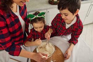 Top view of charming multinational family- loving mother and two cute children kneading dough for Christmas cake in home kitchen. Mom, daughter and son having fun together preparing Christmas cookies photo