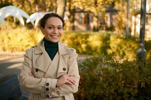 Vertical portrait of attractive business woman in casual beige trench coat and green pullover smiling looking at the camera, posing with crossed arms against the background of autumn nature in a park photo