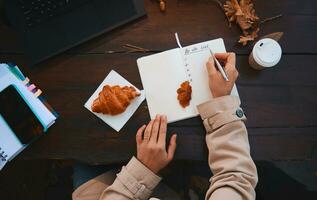 Top view of hand of a cropped woman holding silver pen and writing the list to do in notepad, while sitting at a wooden table with fallen dry oak leaves, takeaway paper cup and baked croissant photo