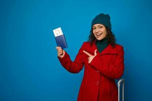 Young smiling African woman in bright red coat, green warm woolen hat, points on passport with air ticket and boarding pass in hands. Travel, air flight journey concept on blue background, copy space photo