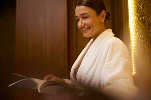 de cerca de un medio Envejecido 30- 40 años antiguo elegante europeo mujer, atractivo morena vestido en blanco terry bata de baño, sonrisas con dientes sonrisa leyendo revista mientras descansando a bienestar spa salón zona foto