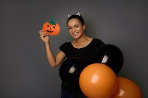 Young pretty woman wearing hoop with cat ears, dressed in black attire, holding colorful black and orange air balloons and shows a felt-cut handmade pumpkin, smiles toothy smile looking at camera photo