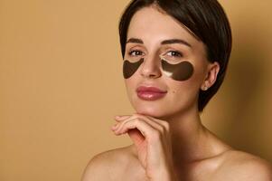 Headshot of beautiful dark-haired woman with shiny healthy clean skin wearing smoothing collagen patches under eyes, isolated over beige background with copy space. Skin care, beauty treatment concept photo