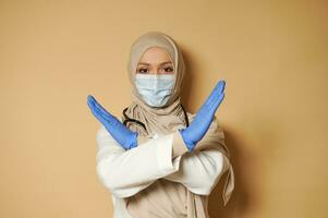 Woman in hijab and medical mask gesturing expressing denial and refuse on beige background with copy space photo