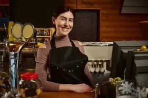 joven atractivo sonriente barista en delantal en pie detrás el bar foto