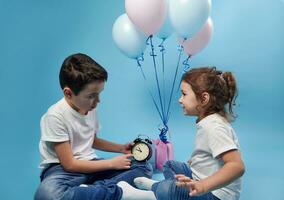 Little boy holding an alarm clock and showing it to his sister on the background of colored balloons. photo