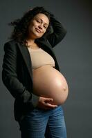Happy curly multi-ethnic pregnant woman gently holding her naked belly, smiling looking at camera, isolated on gray photo