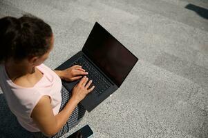 Top view of a woman sitting on steps and typing text on on laptop with blank black monitor screen with copy space for advertising on the urban background. Business concept photo