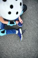 High angle view of boy in safety helmet for skateboard tying shoelaces sitting on asphalt of playground photo