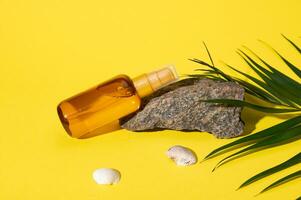 Still life with mockup bottle of sunscreen lotion on a small rock, sea shells and palm leaf, isolated yellow background. photo