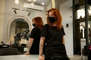 Attractive red-haired woman professional hairdresser in black uniform wearing safety black medical mask looking aside leaning on a countertop in front of a mirror in luxury beauty salon barbershop photo