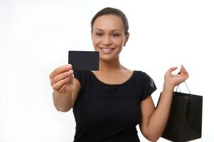 Focus on blank plastic black credit discount card with copy space in outstretched hand of beautiful smiling woman dressed in black and holding shopping bag, isolated on white background. Black Friday photo