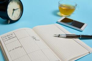 An open agenda with schedule, smartphone, ink pen, glass cup of tea and black alarm clock lie on blue surface. Color background with copy space . Time management, deadline, schedule concept photo