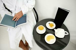 Cropped man in white bathrobe reading magazine while relaxing sitting near served table with lemon slices on a saucer, bowl with honey, teacup and teapot with herbal tea in privacy room of spa center photo