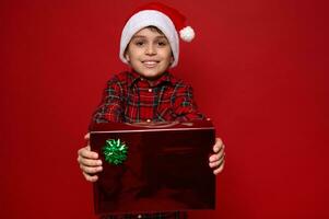 Charming young boy in Santa hat and red plaid shirt holds a Christmas gift in red glitter wrapping paper with shiny green bow in his outstretched hands and showing it to the camera. Copy space for ad photo