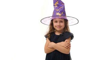 Portrait of winsome little witch smiling girl wearing a wizard hat and dressed in stylish carnival dress, looking at camera posing with crossed arms against white background, copy space, Halloween photo