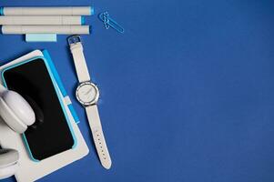 Flat lay of headphones, smartphone in blue case on white organizer, a hand watch, markers and paper clips isolated on blue background with copy space. Top view of white and blue office school supplies photo