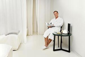 Confident portrait of a handsome mature Caucasian man looking at camera, sitting on an armchair with a magazine in his hands while relaxing in private room of a luxurious wellness spa centre photo