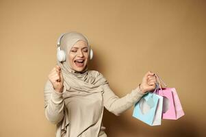 Muslim woman in hijab with headphones holding colored paper bags in her hands enjoying the upcoming religious holidays photo