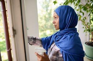 Close-up of confident young Arab Muslim pretty woman with covered head in blue hijab washing windows, removing stains and wiping with rag during spring cleaning in house. Copy space for advertisement photo